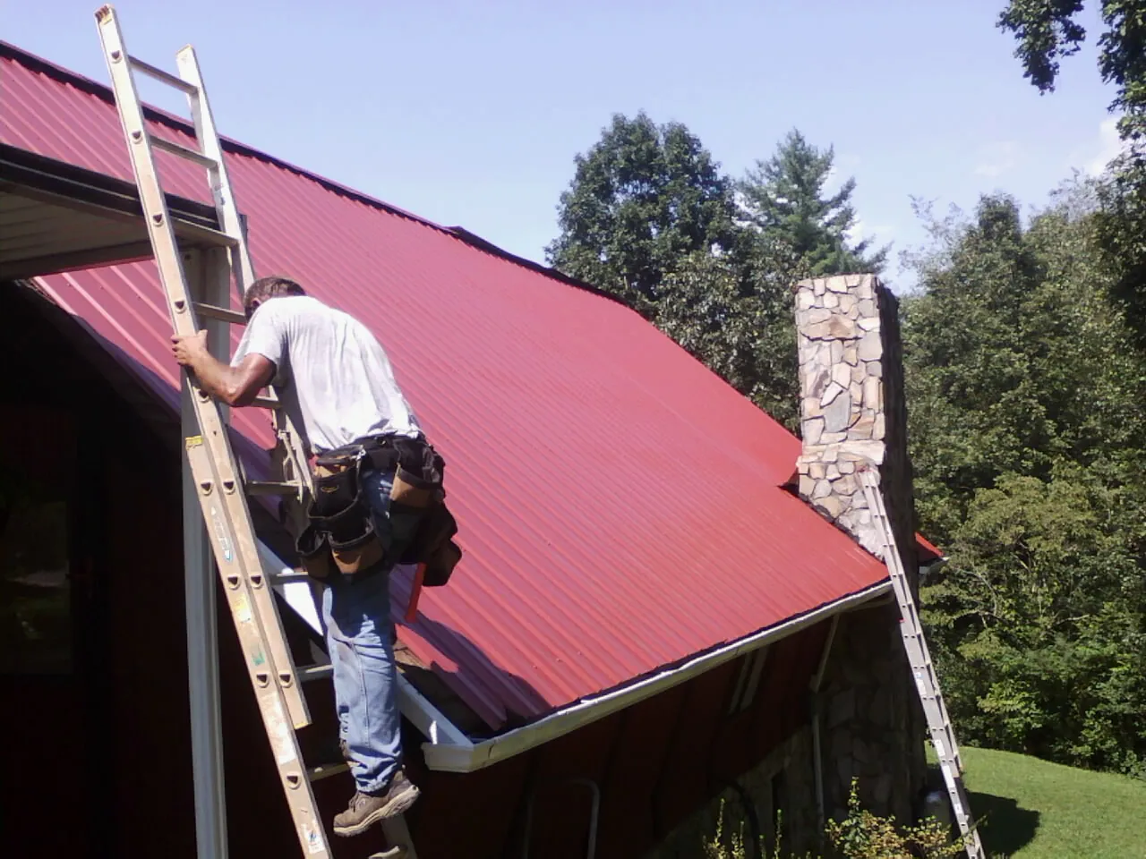 Roofer on a ladder beside a red metal roof and stone chimney — Soffit & Fascia Repair by Roofing Brothers in Chino